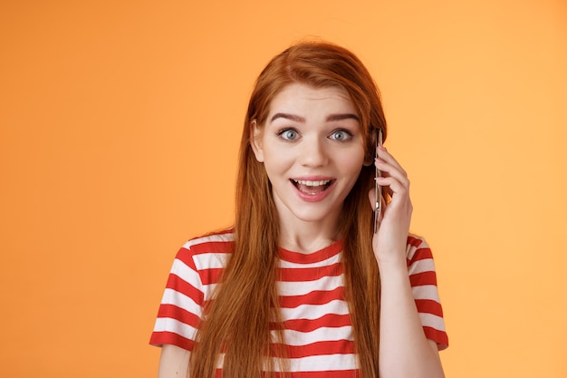 Happy hear friend voice, calling abroad. Amused surprised cute redhead cheerful girl pick up phone, hold smartphone ear, find out awesome news, smiling amazed, stand orange background excited