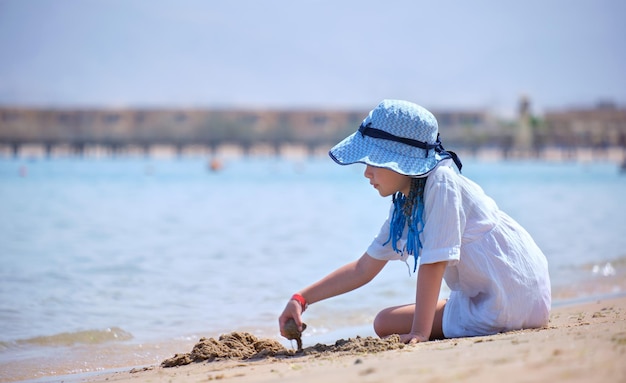 Happy hild girl in big hat and white dress playing alone with wet sand on sandy beach near clear sea lagoon water