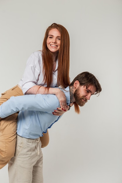 Happy hipster man carrying his girlfriend on his back against a gray background Beautiful funny fashion couple