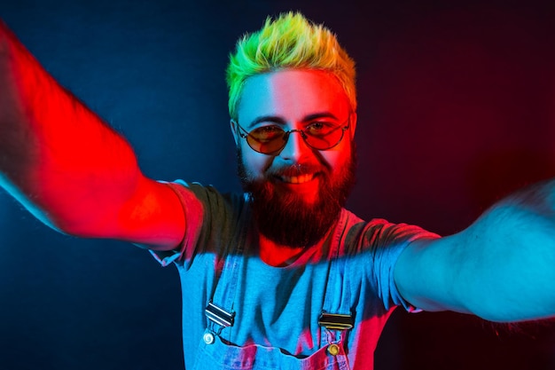 Happy hipster man with pleasant appearance in stylish denim overalls looking at selfie camera with toothy smile, sharing positive with followers. Colorful neon light, indoor studio shot.