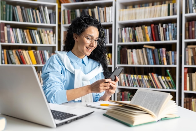 Happy hispanic latino student using her smartphone in a library education technology and learning