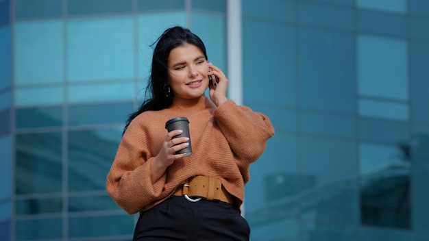 Happy hispanic woman standing outdoors drinking coffee of paper cup responding to friendly call