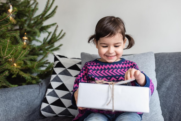 Happy holidays and Merry Christmas Cute little toddler girl with christmas present Kid enjoy the holiday and open the gift sitting on grey sofa in the room