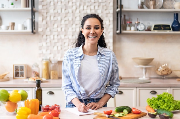 Happy housewife writing new food recipes in her notebook standing in home kitchen interior and