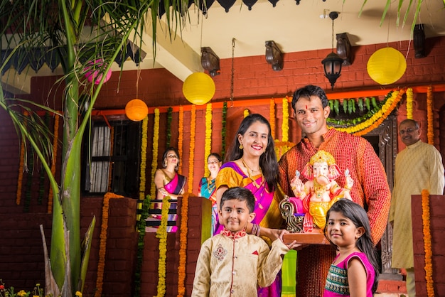 Happy Indian Family Celebrating Ganesh Festival or Chaturthi - Welcoming or performing Pooja and eating sweets in traditional wear at home decorated with Marigold Flowers