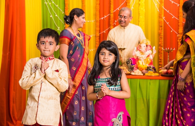 Happy Indian Family Celebrating Ganesh Festival or Chaturthi - Welcoming or performing Pooja and eating sweets in traditional wear at home decorated with Marigold Flowers