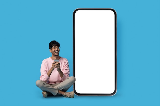 Happy indian guy hipster sitting by big smartphone mockup