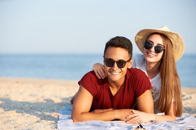 Happy interracial couple newlyweds lie in the sun on tropical travel location Boyfriend and girlfriend relax at spa resort sunbathing near blue lagoon Woman with teeth braces wearing straw hat