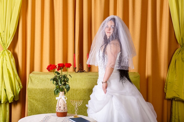 A happy Jewish bride in a fluffy white dress, her face covered with a veil, stands in the room at the table before the chupa ceremony