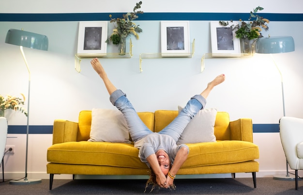 Happy and joyful adult woman lay down on the sofa in reverse position with legs up and head down smiling and having fun at home - middle age young lady active lifestyle laughing a lot on the couch