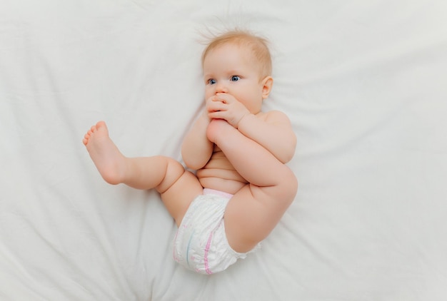 A happy joyful baby in diapers is lying on a white bed and holding his leg Highquality photo place for text