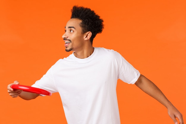 Happy, joyful charismatic african-american man playing with friends, throwing red frisbee left and smiling, spending time outdoors in park, having picnic, vacation over orange wall