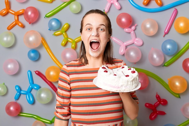 Photo happy joyful cheerful woman with brown hair wearing striped dress holding cake standing against gray wall decorated with colorful balloons posing with sweet dessert on her face screaming with joy