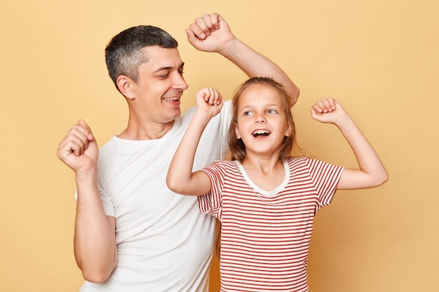 Happy joyful family father and daughter wearing casual tshirts standing isolated over beige background dancing having fun together smiling happily