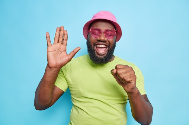 Happy joyful man raises palm sings song keeps hand near mouth as if microphone has upbeat mood wears casual green t shirt pink panama trendy sunglasses isolated over blue wall