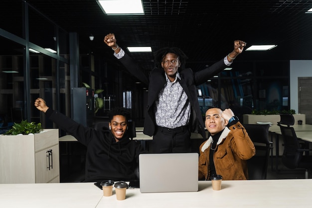 Happy and joyful multiethnic men raise their hands and fists and rejoice in winning in online casino on laptop Teambuilding of black Africans and Asian programmers