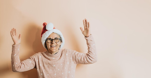 Happy Joyful senior old white hair woman wearing santa claus hat smiling to camera playing. celebrating christmas. stands against a pastel background.Christmas clothes. Copy space. Advertising space