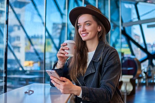 Happy joyful stylish hipster casual woman in hat with mobile phone holds paper cup during drinking coffee and relaxing in coffee cafe. Modern people lifestyle