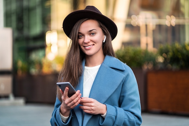 Happy joyful stylish trendy brunette hipster woman in hat and in a blue coat with smartphone and wireless white headphones enjoys and listens music in the city centre. Modern people and technology