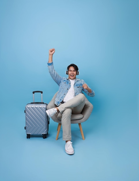 Happy joyful young Asian tourist man listening to music raising his arm dancing sitting on chair