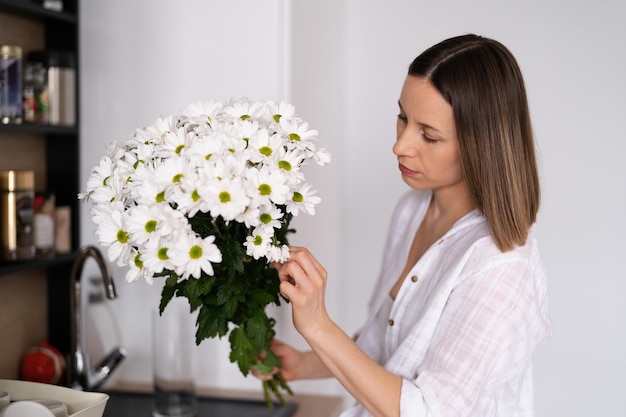 Happy and joyful young woman in white arranging white flowers at home in the kitchen