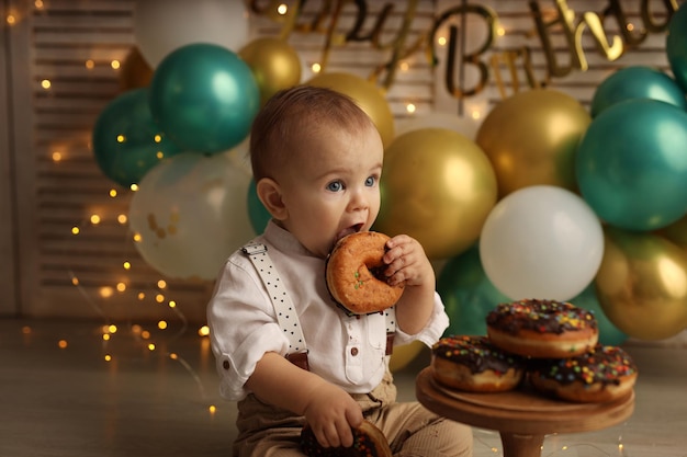 A happy kid on the background of balloons with garlands eats chocolate donuts