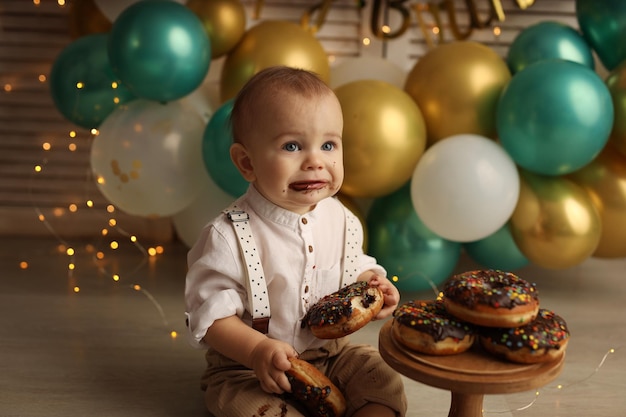 A happy kid on the background of balloons with garlands eats chocolate donuts