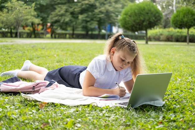 happy kid girl on school uniform with notebook has online lesson on laptop on lawn