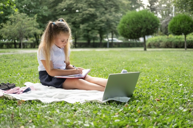 happy kid girl on school uniform with notebook has online lesson on laptop on lawn