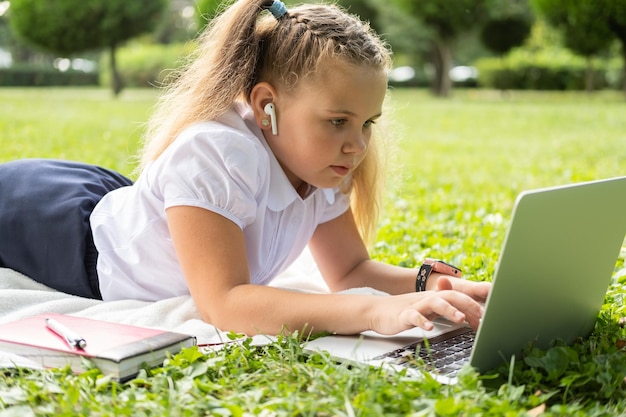 happy kid girl on school uniform with wireless headphone has online lesson on laptop on lawn