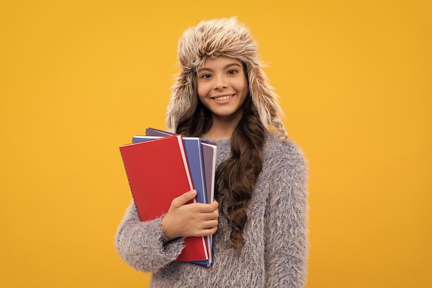 Happy kid long hair in hat with copybook on yellow background education