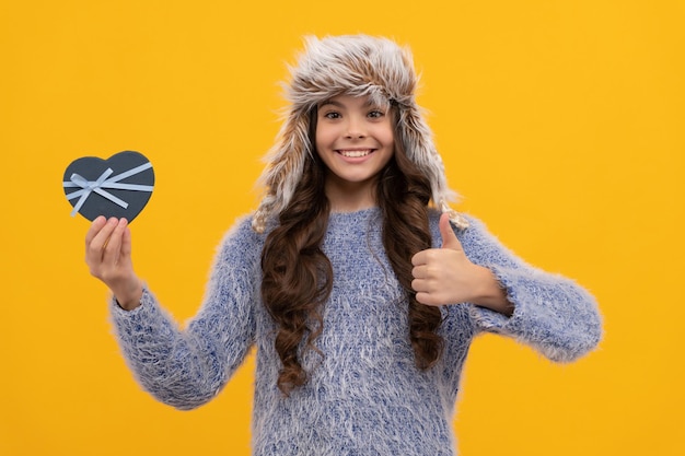 Happy kid long hair in hat with gift box showing thumb up on yellow background valentine