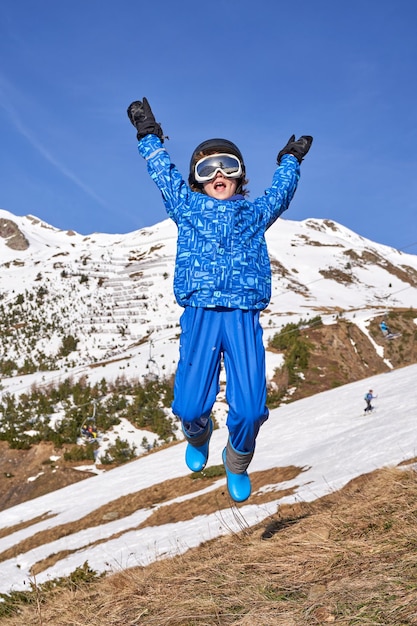 Happy kid in outerwear with goggles and helmet raising arms and screaming while jumping on slope against snowy mountain and blue sky on weekend day on ski resort