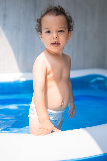 Happy kid playing in an inflatable pool in the backyard Curly toddler smiling and playing with water