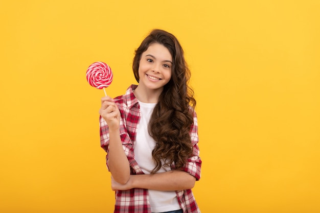 Happy kid with long curly hair in checkered shirt hold lollipop caramel candy on yellow background, sweet tooth.