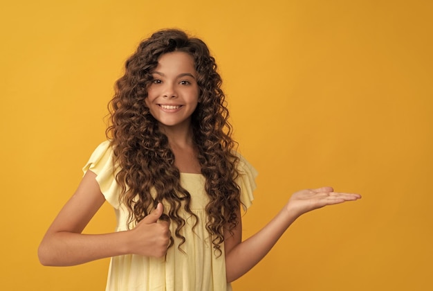 Happy kid with long curly hair and perfect skin presenting product show thumb up copy space advertisement