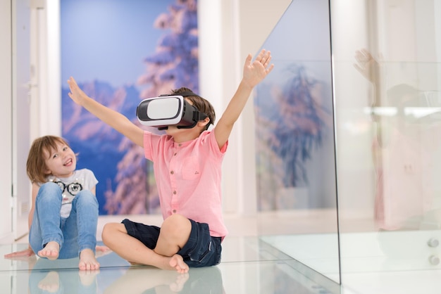 Happy kids using virtual reality headsets and pointing up with finger while sitting on the floor at modern bright home interior