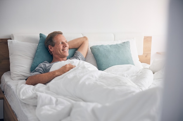 Happy kind adult male person keeping smile on his face while looking aside at window in his bedroom