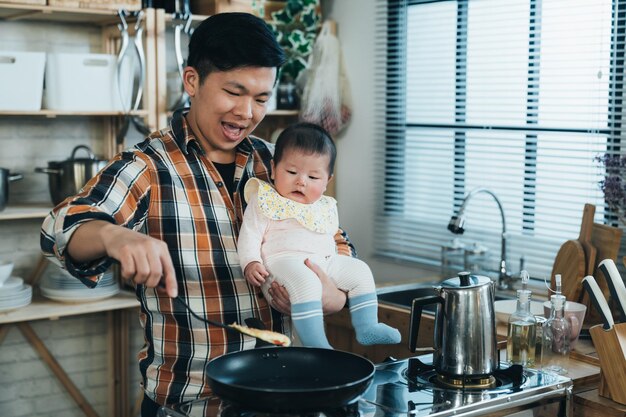 happy Korean father carrying his innocent baby girl in arm is using a spatula to fry food by the kitchen stove during daytime at home.