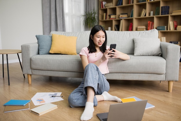 Happy korean lady relaxing at home with laptop and smartphone sitting in living room and messaging on mobile phone