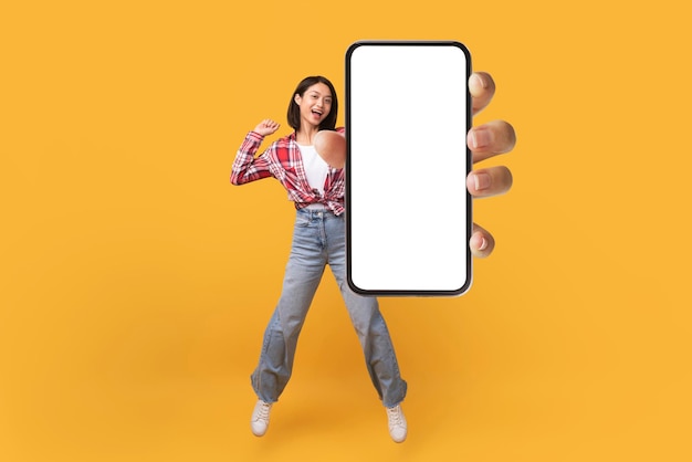 Happy korean lady showing empty smartphone screen while jumping up over yellow studio background collage mockup