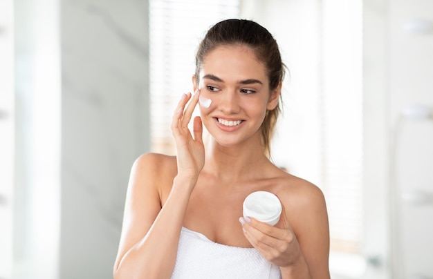 Happy lady applying facial cream holding moisturizer jar in bathroom