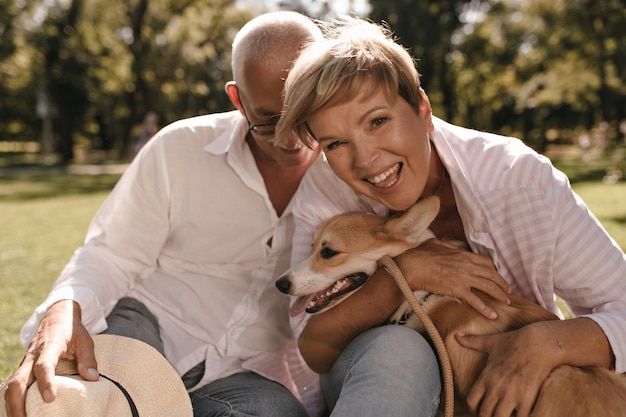 Happy lady with short hairstyle in pink shirt laughing hugging dog and posing with grey haired man in white long sleeve shirt in park