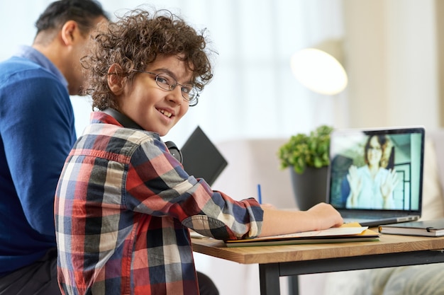 Happy latin boy wearing glasses sitting at the desk together with his father, using laptop while having online lesson with teacher during remote learning at home. Online education, homeschooling