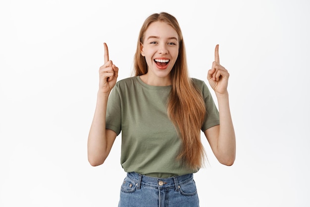 Happy laughing girl with blond hair, pointing and showing advertisement on top, recommending click on link banner, standing against white background.