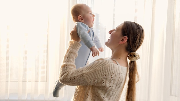 Happy laughing holding and playing with her smiling baby son against sunset in big window Concept of family happiness and parenting