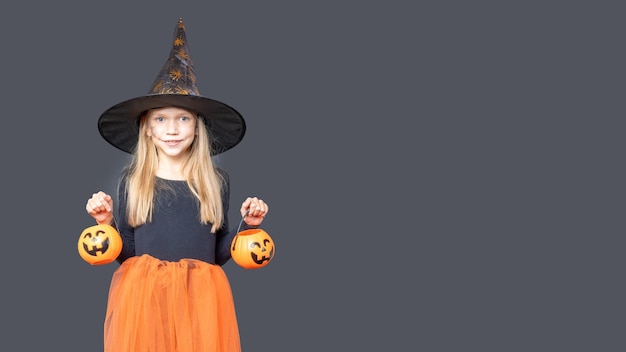 A happy laughing little girl in a carnival witch costume holds a Halloween pumpkin baskets for treats on a dark background