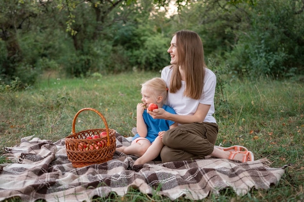 Happy laughing woman with blond daughter hold apple sitting on blanket in park Mother with child had picnic in park