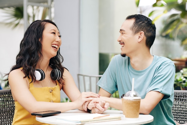 Happy laughing young Chinese couple sitting at cafe table, touching hands and looking at each other