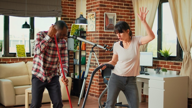 Happy life partners using tools to clean apartment, listening to music and dancing while they use mop and vacuum. Diverse couple cleaning living room floors, sweeping dust with cleaner.